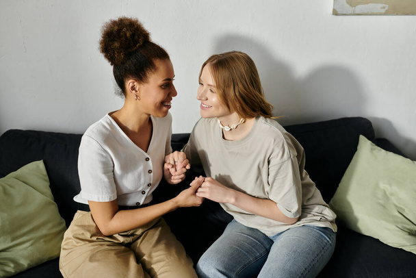 Δύο γυναίκες με καθημερινά ρούχα κάθονται σε έναν καναπέ, κρατώντας τα χέρια και γελώντας.. - Φωτογραφία, εικόνα