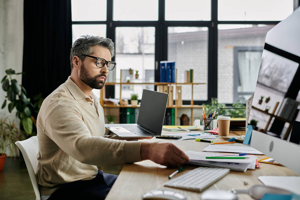 Ein gutaussehender Geschäftsmann mit Bart arbeitet intensiv an seinem Laptop in einem modernen Büroambiente. Papiere und Büromaterial liegen auf seinem Schreibtisch. - Foto, Bild
