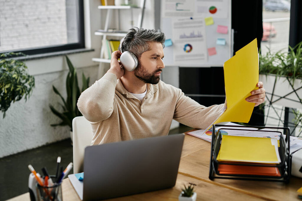 Egy jóképű, szakállas üzletember szorgalmasan dolgozik egy modern irodában, átnézi a dokumentumokat fülhallgató viselése közben.. - Fotó, kép