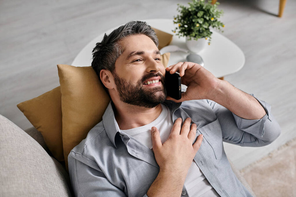 Un hombre barbudo con atuendo casual se sienta en un sofá en casa y se ríe mientras habla por teléfono. - Foto, imagen