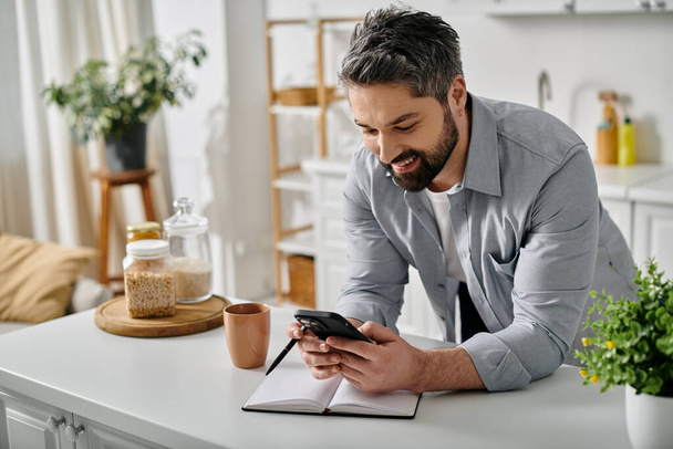 Een man met een baard in een casual shirt werkt op afstand vanuit zijn thuiskeuken, met behulp van zijn telefoon en een notitieboekje. - Foto, afbeelding