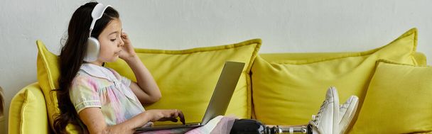 Una joven con una pierna protésica se relaja en un sofá, usando un portátil mientras usa auriculares. - Foto, imagen