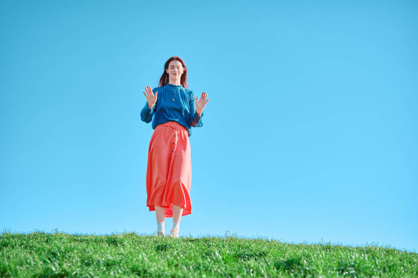 Teen girl in coral skirt standing on green grass against clear blue sky. Summer and casual outfit concept. - Photo, Image