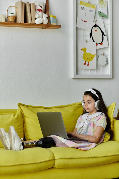 Молодая девушка с протезной ногой сидит на жёлтом диване, используя ноутбук и наушники. Она дома, окружена рисунками на стене, наслаждается временем с технологией. - Фото, изображение