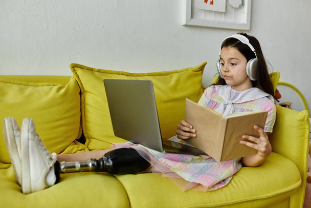 Ένα νεαρό κορίτσι με προσθετικό πόδι κάθεται άνετα σε έναν κίτρινο καναπέ, απορροφημένο στην ανάγνωση ενός βιβλίου.. - Φωτογραφία, εικόνα