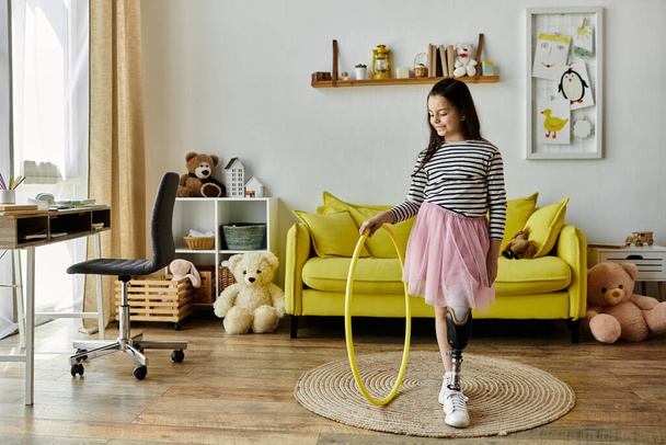 Uma jovem com uma perna protética gira um aro de hula amarelo em sua sala de estar em casa, cercada por brinquedos e um sofá amarelo. - Foto, Imagem