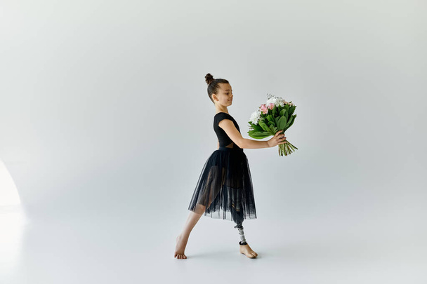 Uma jovem com uma perna protética graciosamente segura um buquê de flores enquanto usa um vestido de balé preto em um estúdio. - Foto, Imagem