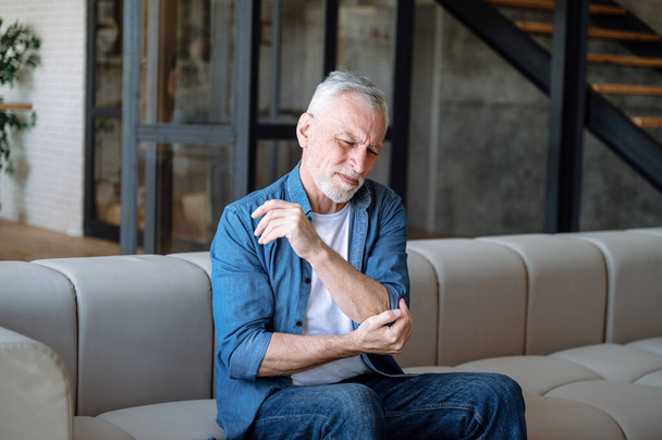 Грустный зрелый мужчина сидит один на диване в квартире, чувствует себя плохо, трогает локоть, страдает от ревматоидного артрита и боли в суставах. Концепция здравоохранения - Фото, изображение