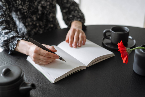 Valikoiva keskittyä muistikirja tyhjiä sivuja musta pöytä teekannu, kuppi ja pieni maljakko kukka. Nainen, jolla on kynä ideoiden kirjoittamiseen paperille. Muistiinpanojen tekeminen muistioon - Valokuva, kuva