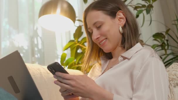 Νεαρή γυναίκα φαίνεται χαρούμενη και χαλαρή, χρησιμοποιώντας smartphone και laptop στο σύγχρονο, φωτεινό χώρο εργασίας στο άνετο, φυτεμένο σπίτι - Πλάνα, βίντεο