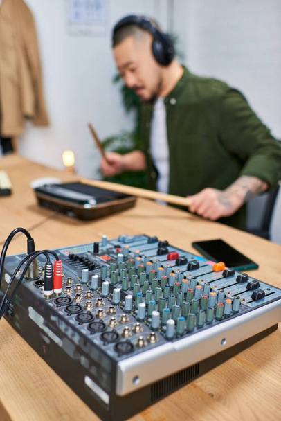 Primer plano de un mezclador de sonido en un estudio con una imagen borrosa de un hombre asiático tocando un instrumento en el fondo. - Foto, Imagen
