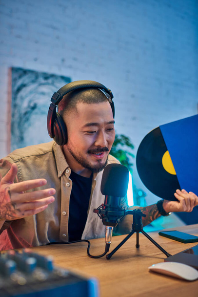 Un joven asiático con auriculares y una camisa beige está grabando un podcast en su estudio. Está hablando en un micrófono y haciendo gestos con sus manos.. - Foto, Imagen