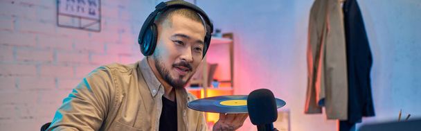 Egy jóképű ázsiai férfi fülhallgatót visel, és lemezt tart a stúdiójában.. - Fotó, kép
