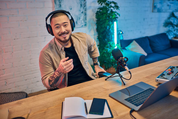 Un uomo asiatico sorridente che indossa le cuffie registra un podcast nel suo studio. - Foto, immagini