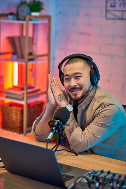 Ένας όμορφος Ασιάτης που φοράει ακουστικά χαμογελά ενώ κάνει podcasting στο στούντιο του σπιτιού του.. - Φωτογραφία, εικόνα