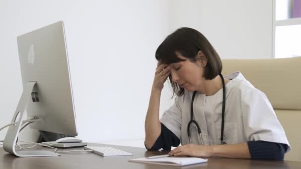 Stressed young female doctor sitting with head in hands at computer desk as she suffers from headache while being overworked in clinic - Footage, Video