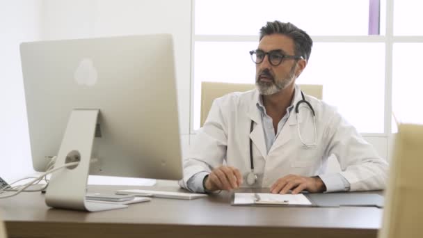 Felice maturo medico maschio con stetoscopio intorno al collo utilizzando PC desktop e guardando la fotocamera mentre seduto sulla sedia in clinica - Filmati, video