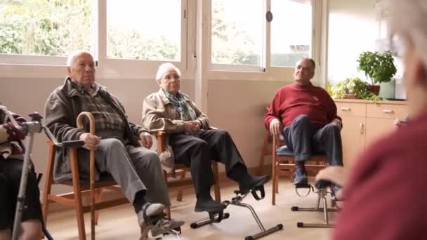 Anziani seduti su sedie e gambe di allenamento con pedali sotto la scrivania in bicicletta durante la giornata di sole in casa di vecchiaia - Filmati, video