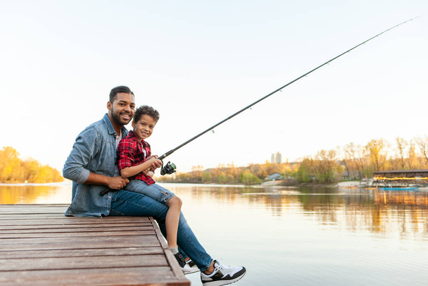 Αφροαμερικάνος και παιδί κάθονται σε μια ξύλινη προβλήτα κρατώντας καλάμια ψαρέματος στο ποτάμι, ο μπαμπάς μαθαίνει στο γιο του να ψαρεύει στη λίμνη, να ξεκουράζεται και να χαλαρώνει το Σαββατοκύριακο. - Φωτογραφία, εικόνα