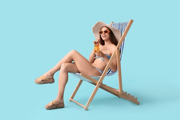 Krásná mladá žena ve stylové pruhované plavky se sklenkou koktejlu sedí v palubní židli na modrém pozadí - Fotografie, Obrázek