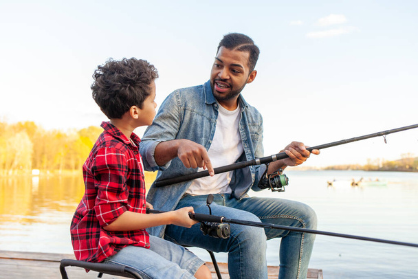 Afroamerykanin i dziecko siedzą na drewnianym pomoście trzymając wędki na rzece, tata uczy syna łowić ryby w jeziorze, rodzina odpoczywa i relaksuje się w weekend - Zdjęcie, obraz