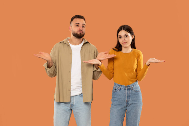 Egy férfi és egy nő egymás mellett állnak, és felemelik a kezüket a zavarodottság gesztusával. Ők öltözött alkalmi, narancs stúdió háttér - Fotó, kép
