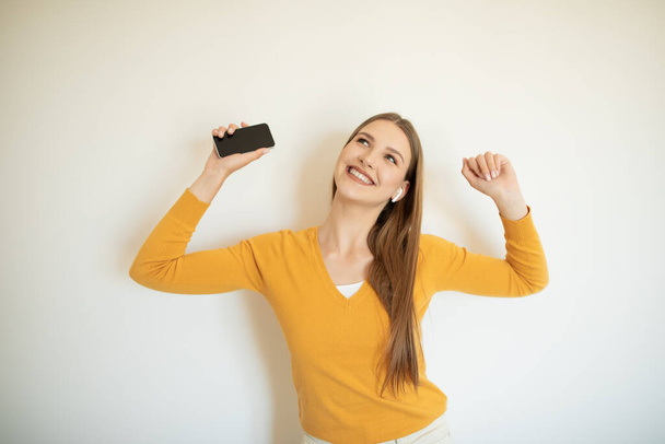 Una adolescente con un suéter amarillo está bailando en el interior, sosteniendo un teléfono inteligente en una mano y levantando su otra mano alegremente. Tiene una gran sonrisa en la cara.. - Foto, imagen