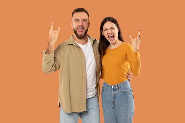 Ein fröhliches Paar macht begeistert rockige Handgesten und lächelt dabei breit. Der Hintergrund ist eine leuchtend orangefarbene Farbe, die ihren fröhlichen Ausdruck und ihre lässige Kleidung hervorhebt. - Foto, Bild