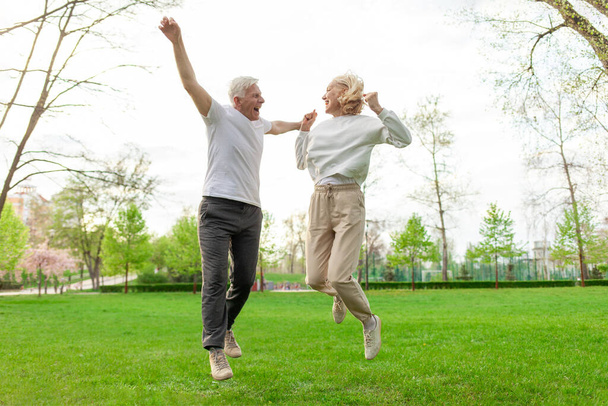 elderly senior couple man and woman rejoice in victory and jump in the park outdoors, gray-haired grandparents celebrate luck and success and rejoice in nature - Photo, Image