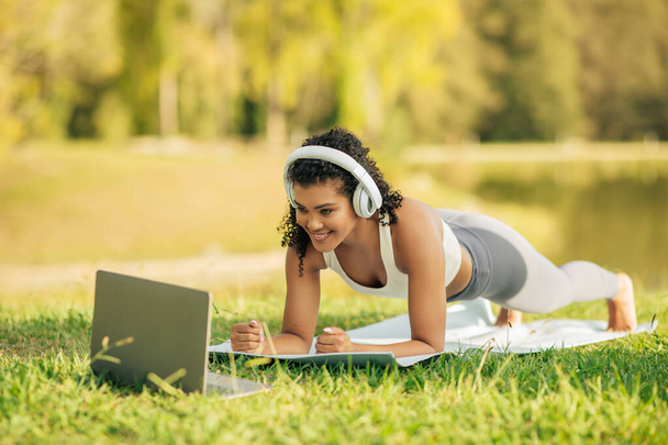 Egy atlétikai ruhás nőt láttak, amint egy jóga szőnyegen gyakorlatozik, miközben laptopot néz, fejhallgatóval a napfényes parkban.. - Fotó, kép