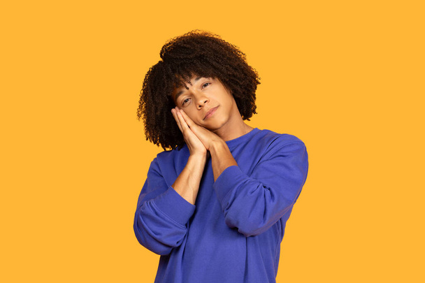 Un giovane con i capelli ricci indossa un maglione blu e posa come se stesse dormendo, con le mani unite sotto la guancia. Si trova di fronte a uno sfondo arancione vibrante - Foto, immagini