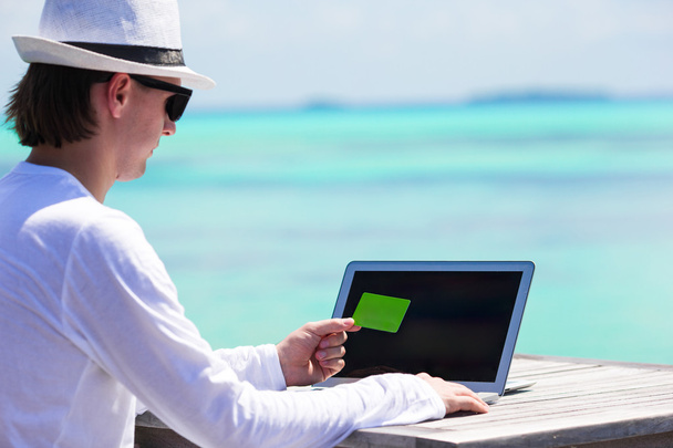 Молодой человек работает на ноутбуке с кредитной картой на тропическом пляже
 - Фото, изображение
