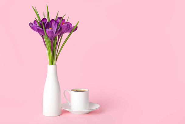 Florero con hermosas flores de azafrán y taza de café sobre fondo rosa - Foto, Imagen