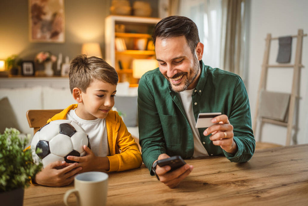 Papá tiene tarjeta de crédito y el hijo tiene pelota de fútbol sentarse en la cocina tienen compras en línea en el teléfono celular - Foto, imagen