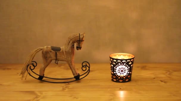 Christmas decoration - wooden toy rocking-horse and candle in candlestick on wooden table against concrete wall - Footage, Video