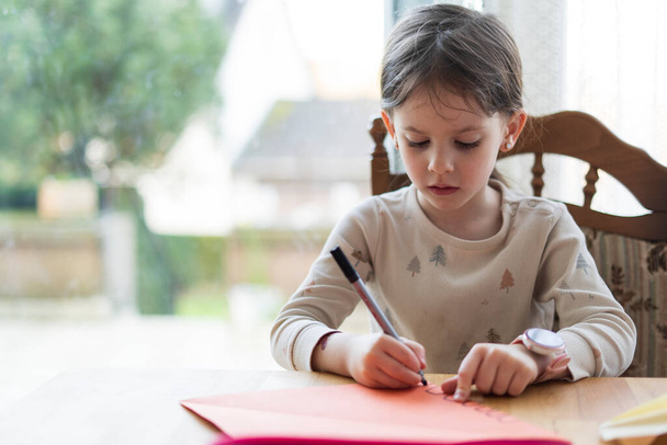 Een schattig meisje met een paardenstaart, zittend aan een zittafel, doet haar huiswerk, houdt een pen in haar hand. Begrip thuisonderwijs. - Foto, afbeelding