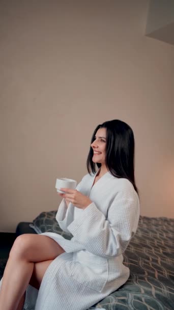 A young woman in a bathrobe drinks coffee and looks out the window of a hotel in the mountains. Hotel holiday concept. Vertical video - Footage, Video