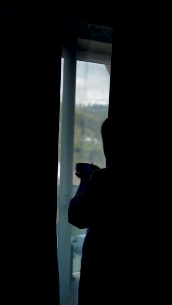 バスローブの若い女性がコーヒーを飲んで,山のホテルの窓を眺める. ホテルホリデーコンセプト。 バーティカルビデオ - 映像、動画