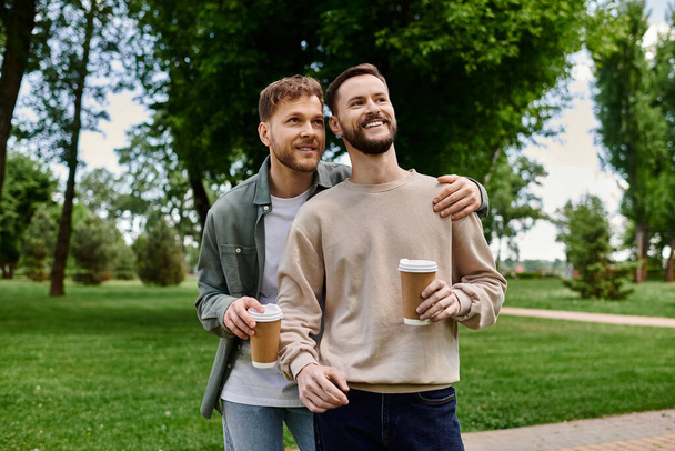 Günlük kıyafetlerle sakallı eşcinsel bir çift parkta yürüyor güneşli bir öğleden sonra ve birlikte kahve içiyorlar.. - Fotoğraf, Görsel