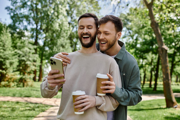 Een bebaarde gay paar deelt een lach terwijl het kijken naar een telefoon en genieten van koffie in een groen park. - Foto, afbeelding