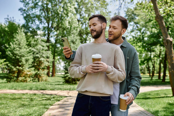 Zwei bärtige Männer in lässiger Kleidung lächeln und schauen auf ihr Handy, während sie durch einen grünen Park spazieren. - Foto, Bild