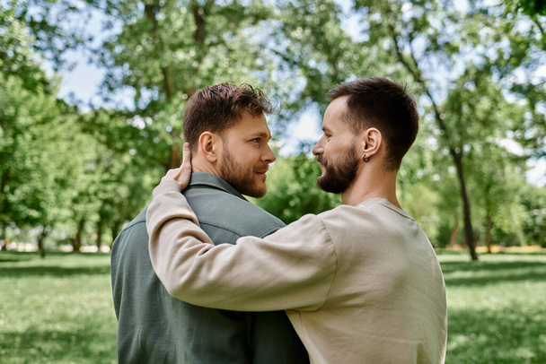 Ein bärtiges homosexuelles Paar steht in einem grünen Park, umarmt und schaut einander in die Augen. - Foto, Bild