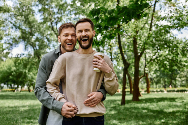 Ein bärtiges schwules Paar in lässiger Kleidung lacht und umarmt sich, während es Zeit miteinander in einem grünen Park verbringt. - Foto, Bild