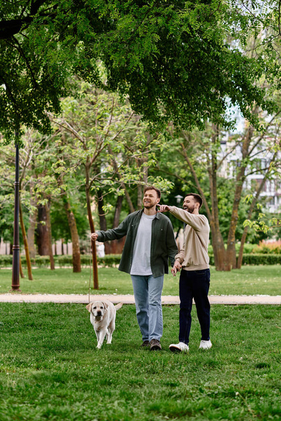 Ein schwules Paar mit Bärten geht Hand in Hand mit seinem Labrador-Retriever-Hund durch einen üppig grünen Park. - Foto, Bild