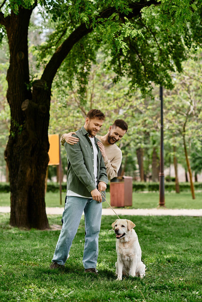 カジュアルな服装のひげ付きゲイカップルは,緑の公園でラブラドールと散歩を楽しんでいます. - 写真・画像