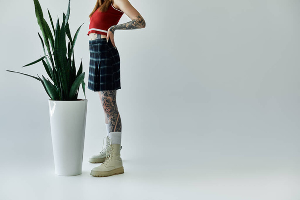 Una giovane donna tatuata indossa una canotta rossa e una gonna a quadri, in piedi davanti a uno sfondo grigio. - Foto, immagini