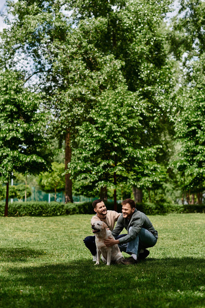 Δύο γενειοφόροι γκέι άντρες με καθημερινά ρούχα απολαμβάνουν μια ηλιόλουστη μέρα σε ένα καταπράσινο πάρκο με το λαμπραντόρ ριτρίβερ τους.. - Φωτογραφία, εικόνα