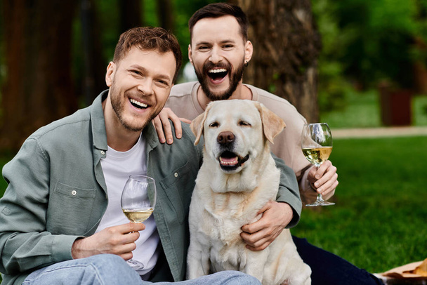 Dos hombres barbudos ríen alegremente con su perro Labrador mientras disfrutan de un picnic en un parque verde. - Foto, Imagen