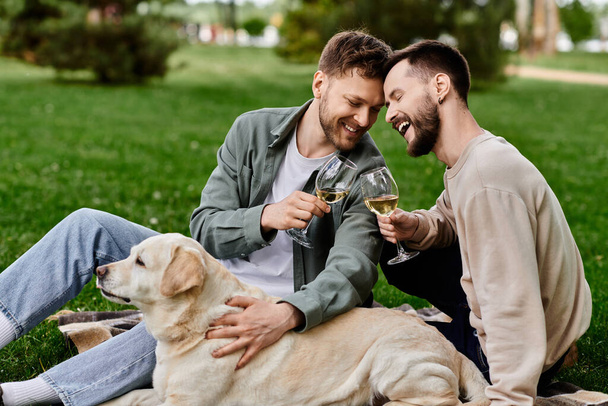 A gay couple enjoys a picnic in the park with their labrador retriever, sharing laughter and wine while enjoying the beautiful green surroundings. - Фото, изображение