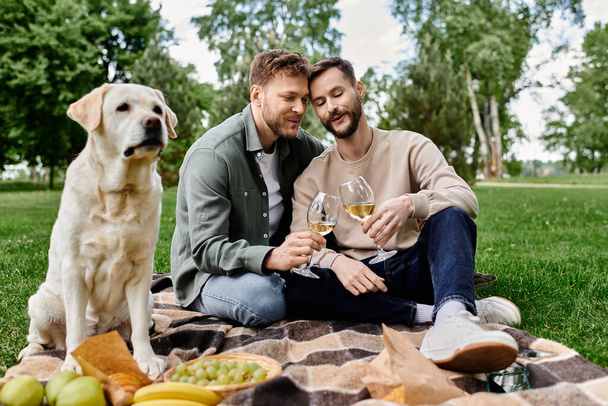 Egy szakállas meleg pár élvezi a pikniket a labrador retrieverével egy buja zöld parkban. Megisznak egy üveg bort, és úgy tűnik, romantikus és pihentető időtöltésük van.. - Fotó, kép